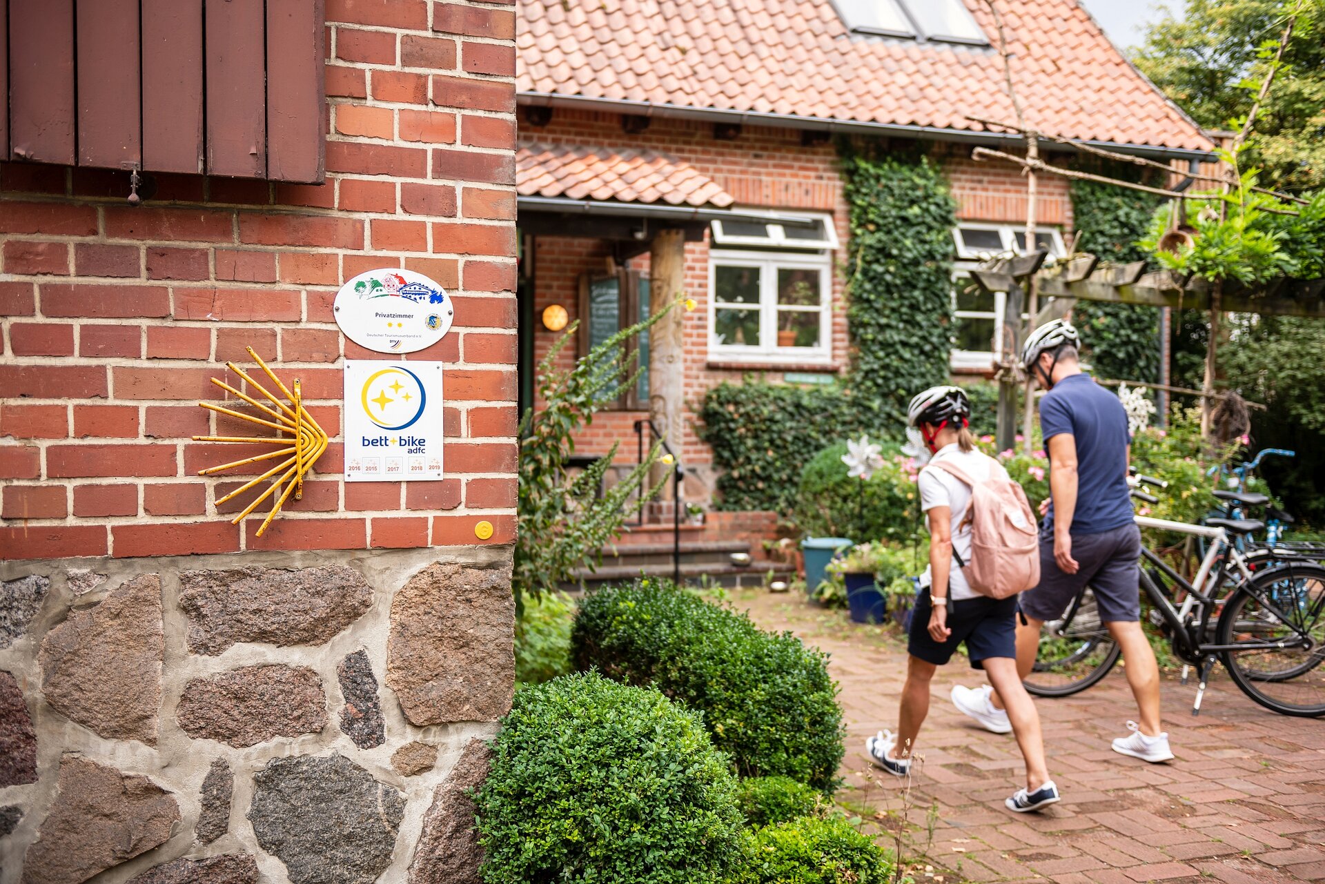 Ein Paar mit Fahrradhelmen geht auf eine Bett+Bike-Unterkunft zu. Im Vordergrund die Bett+Bike-Plakette an der Hauswand.