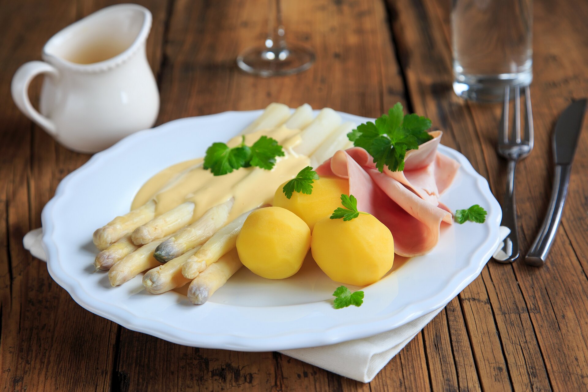 Ein Holztisch mit Essbesteck, einem Kännchen und einem Teller auf dem Spargel, Salzkartoffeln und Schinken angerichtet sind