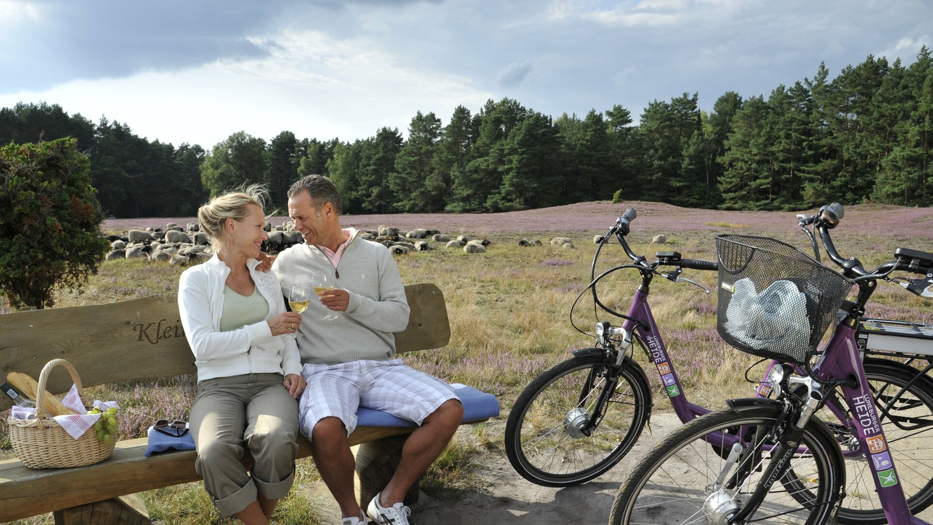 Ein Paar im Seniorenalter sitzt auf einer Bank in der Klein Bünstorfer Heide und trinkt Wein. Neben ihnen stehen zwei Pedelecs.