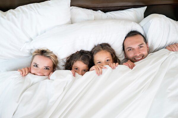 Eine Familie Frau, zwei Kinder und ein Mann, liegt nebeneinander in einem großen Bett. Die weiße Bettdecke haben sie mit den Händen bis unters Kinn gezogen, sodass nur die Köpfe hervorschauen.